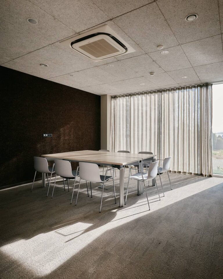 Minimalist meeting room with a square wooden table, white chairs, and natural daylight filtering through sheer curtains