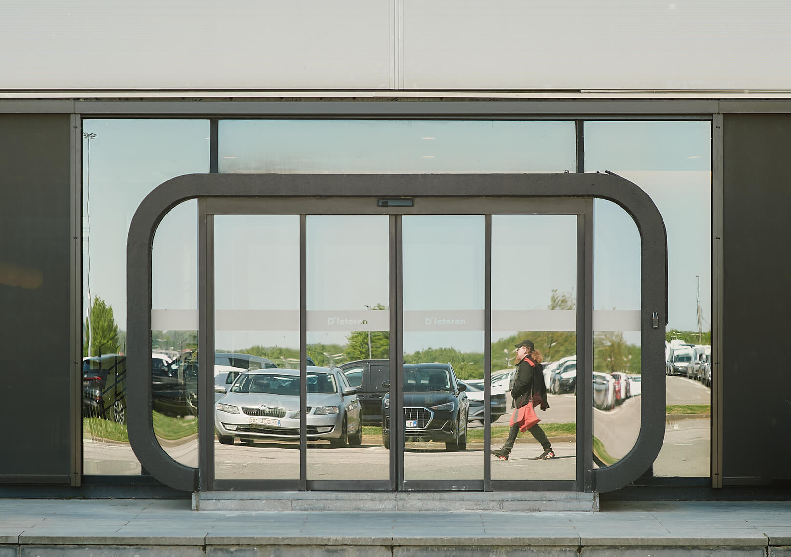Glass entrance of a modern building reflecting parked cars and a person walking by, with the name 'D'Ieteren' visible on the doors