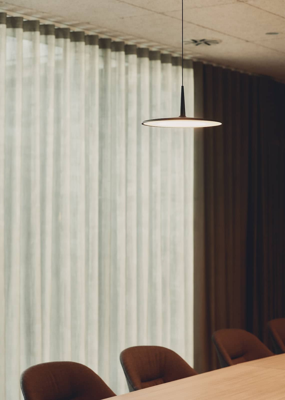 Modern circular pendant light above meeting chairs with layered curtains in the background.
