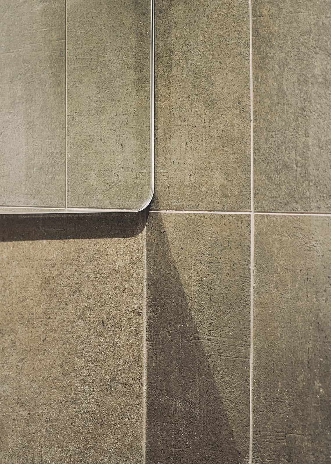 Detail of a textured concrete tile wall with a mirror featuring rounded corners.