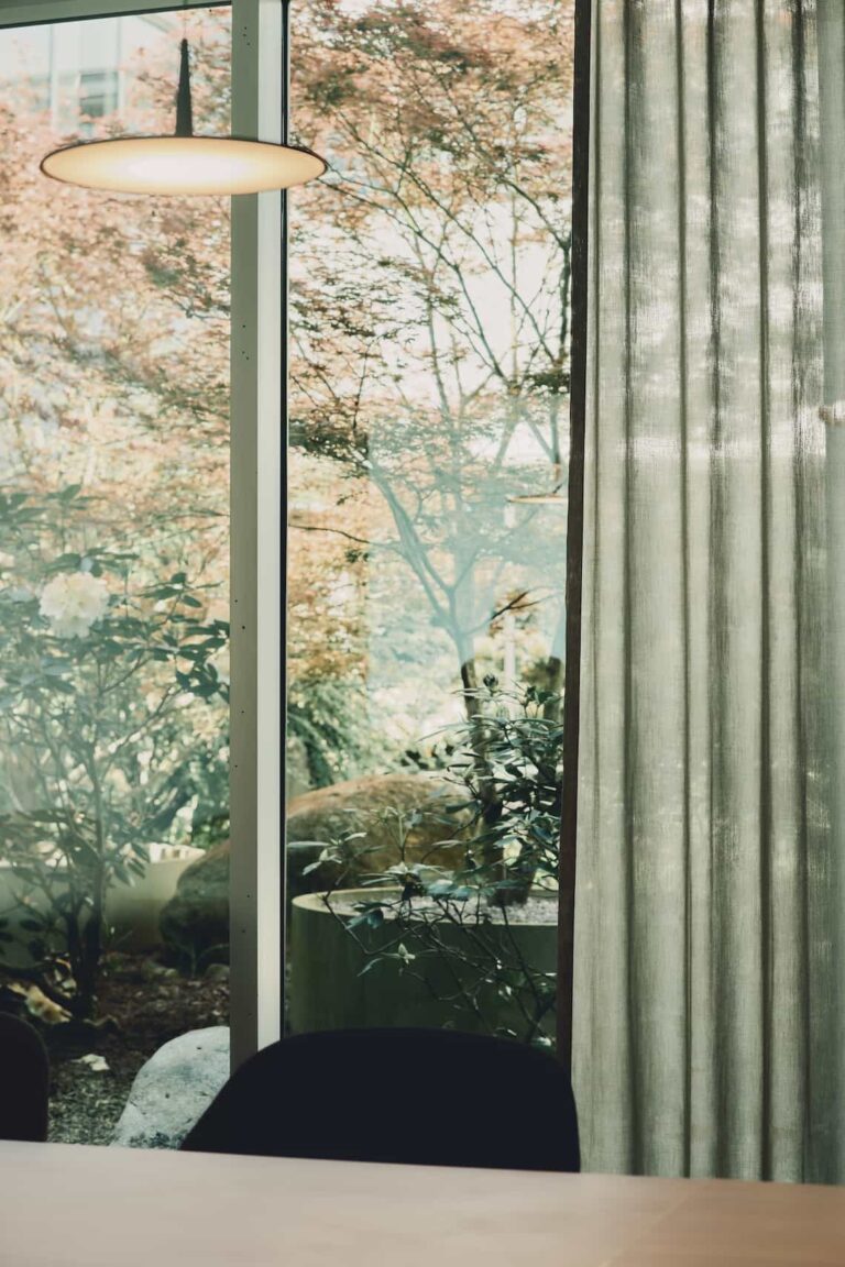 Lush garden with a maple tree and flowering bushes, seen through a glass wall next to a green outdoor seating area.