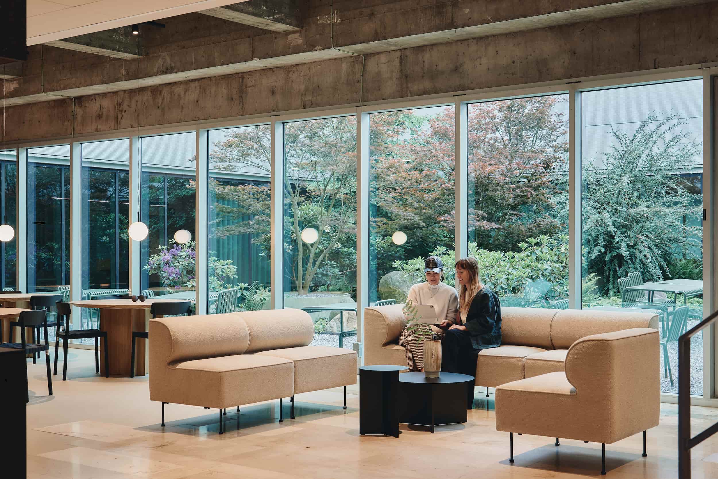 Two people sitting on a modular beige sofa in a lounge, facing large windows with views of a leafy courtyard.