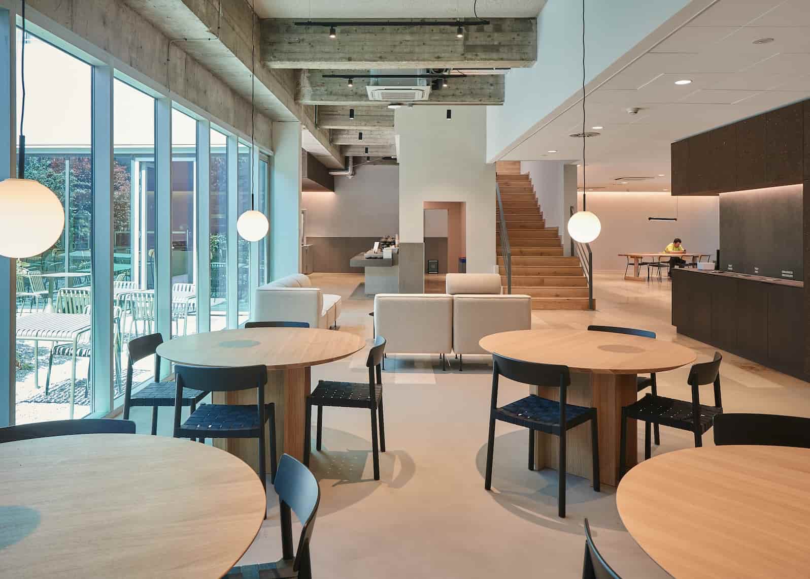 Bright and modern interior lounge with round wooden tables, black chairs, white sofas, spherical pendant lights, and large windows overlooking a garden. A wooden staircase and a person working in the background add depth to the open space.