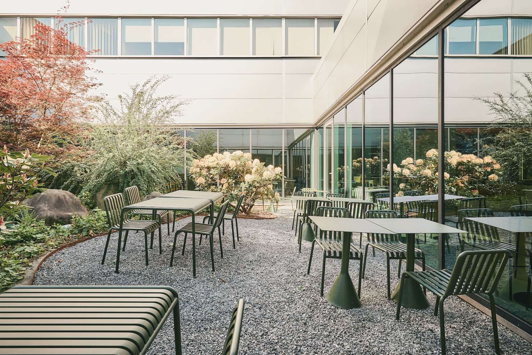 Tranquil garden courtyard with green metal tables and chairs on gravel ground, surrounded by flowering plants and adjacent to a modern building with large glass windows