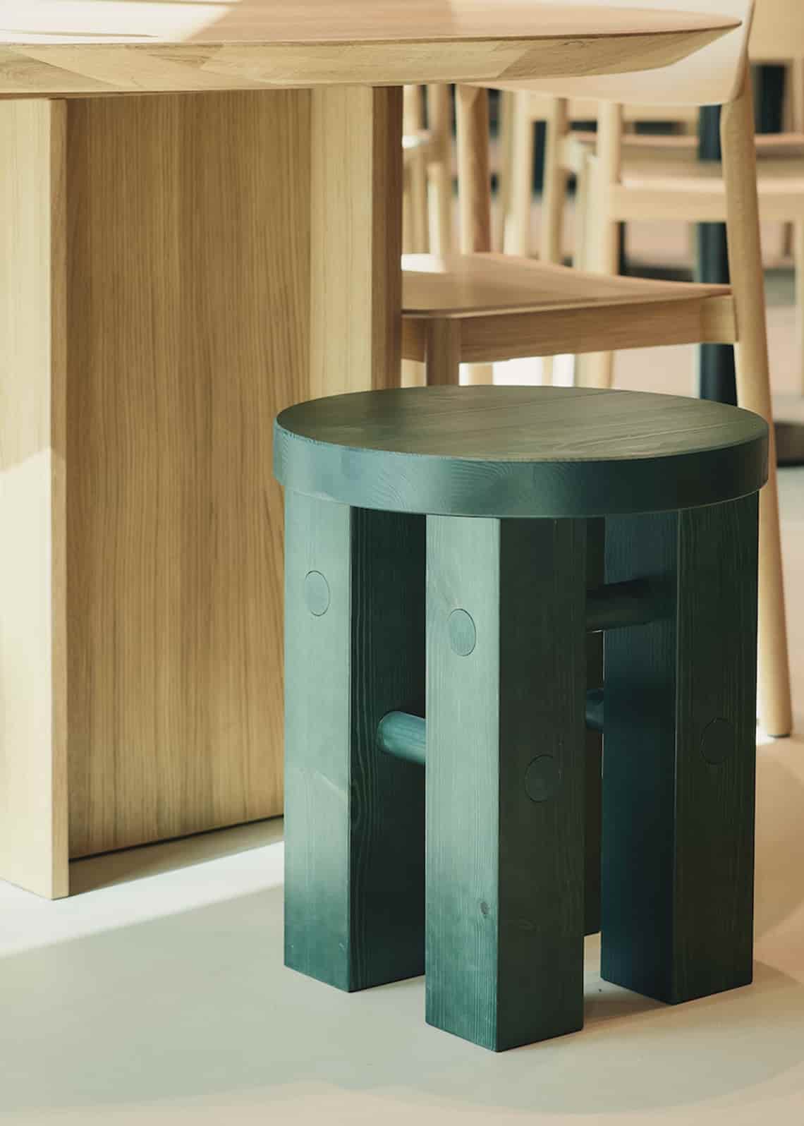 Close-up of a round green wooden stool placed under a light oak table in a modern interior.