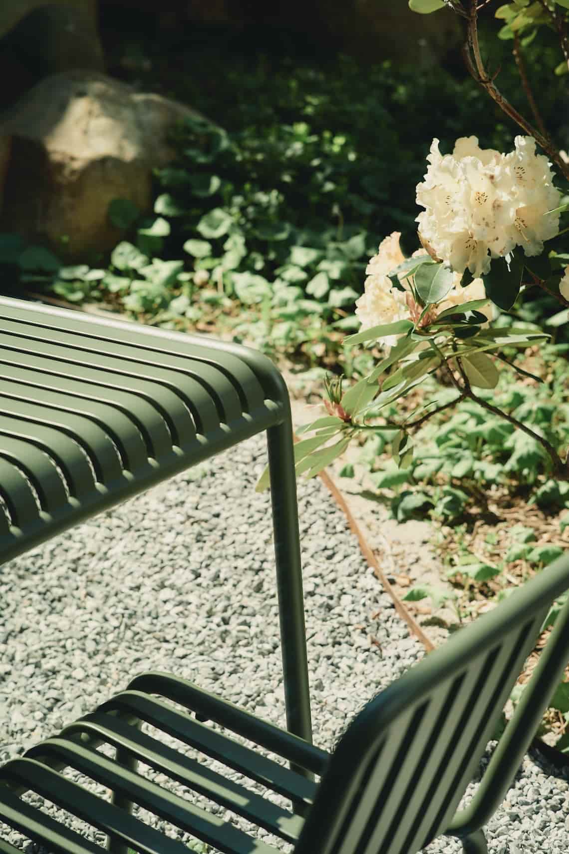 Outdoor green metal chair next to blooming white flowers and gravel flooring, surrounded by greenery.