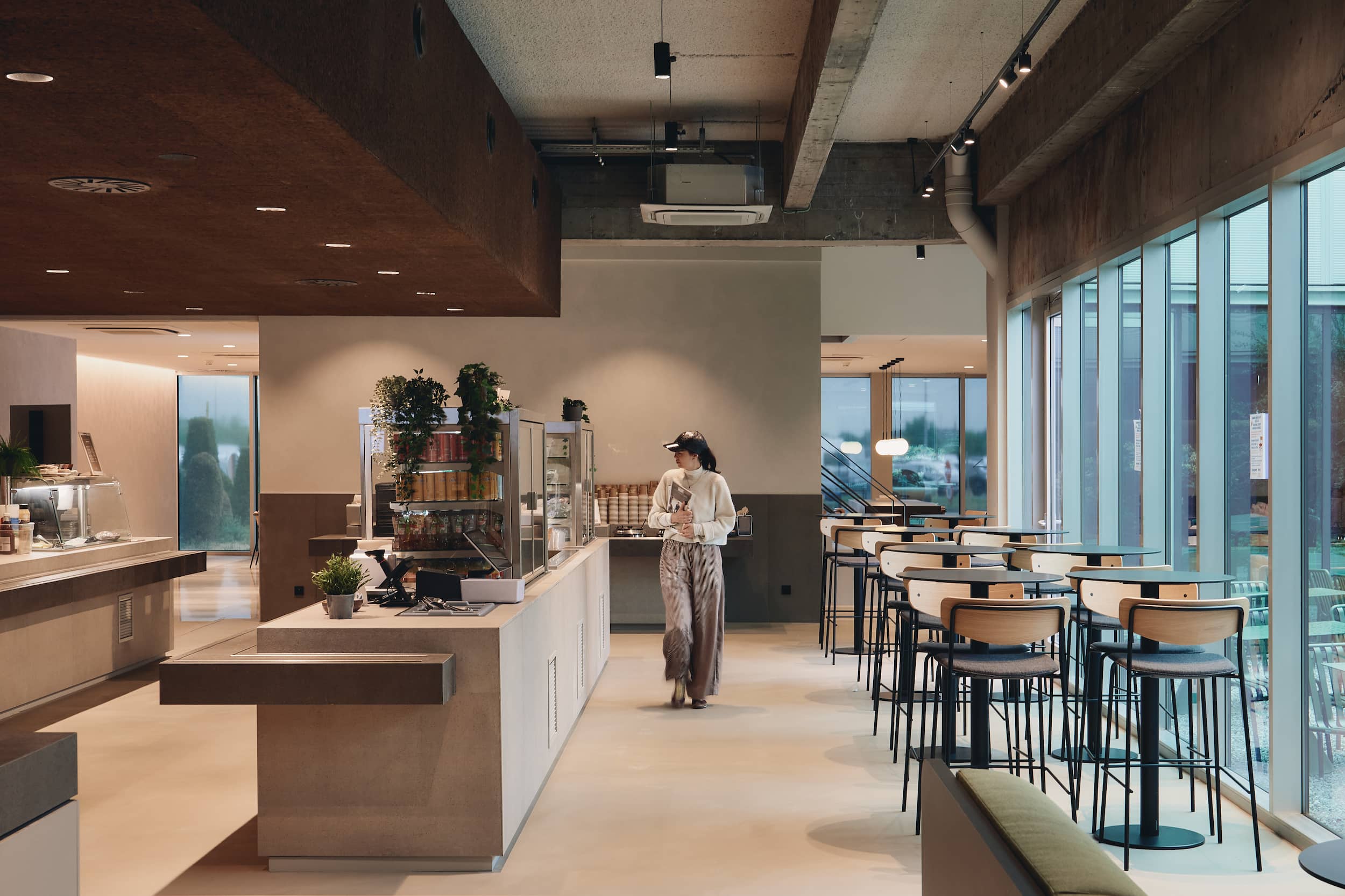 A woman walks through a modern kitchen space with minimalist counters, pendant lighting, and large windows.