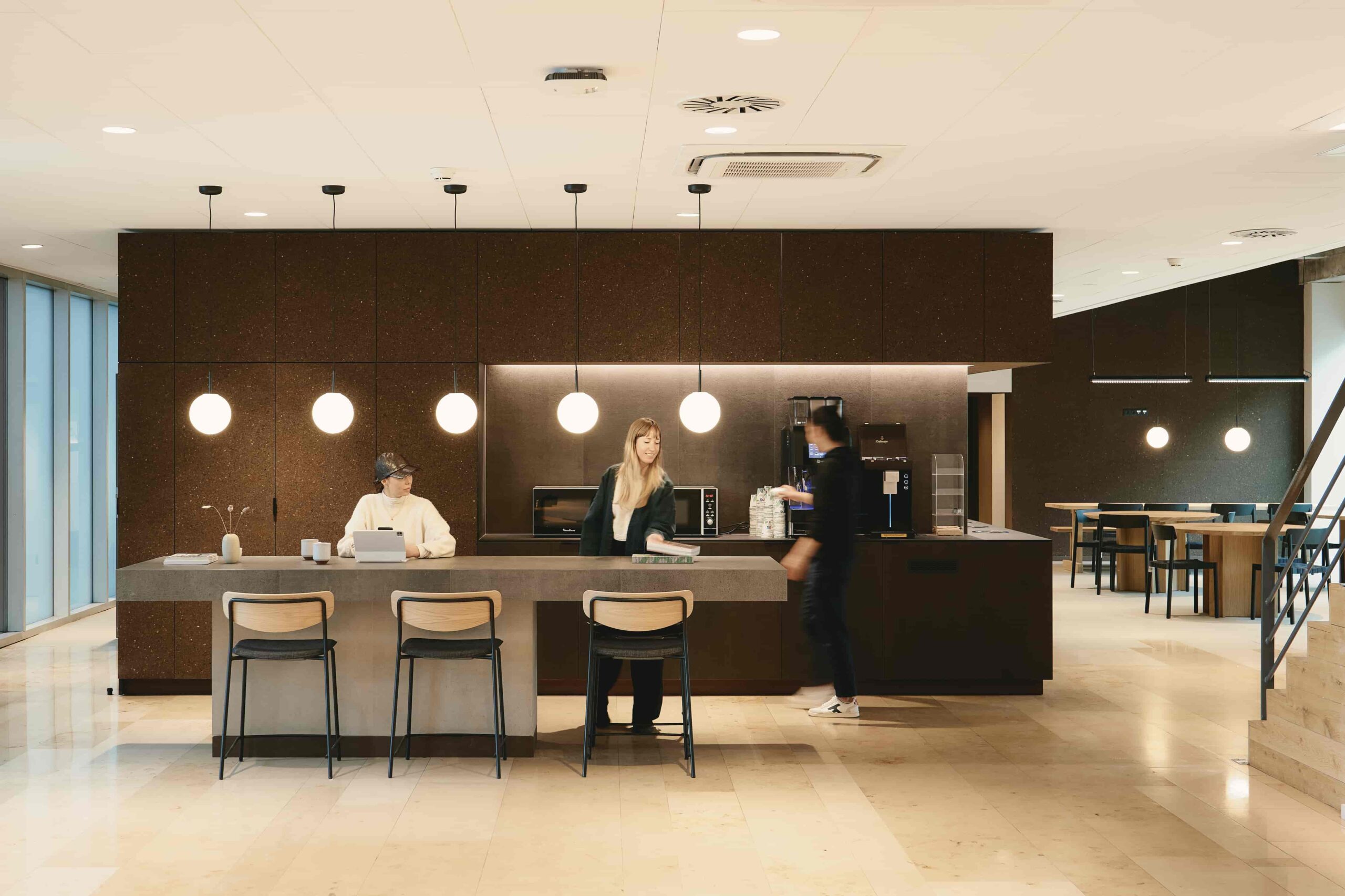 Modern office coffee lounge area with pendant lighting, a central island, coffee machines, and people interacting and working in a relaxed atmosphere