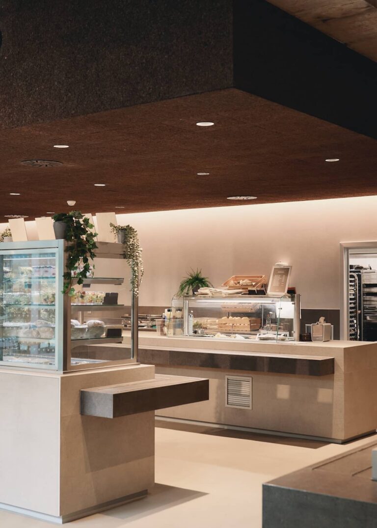 Display counter in a contemporary kitchen featuring built-in glass shelving, potted plants, and an assortment of sandwiches and baked goods.