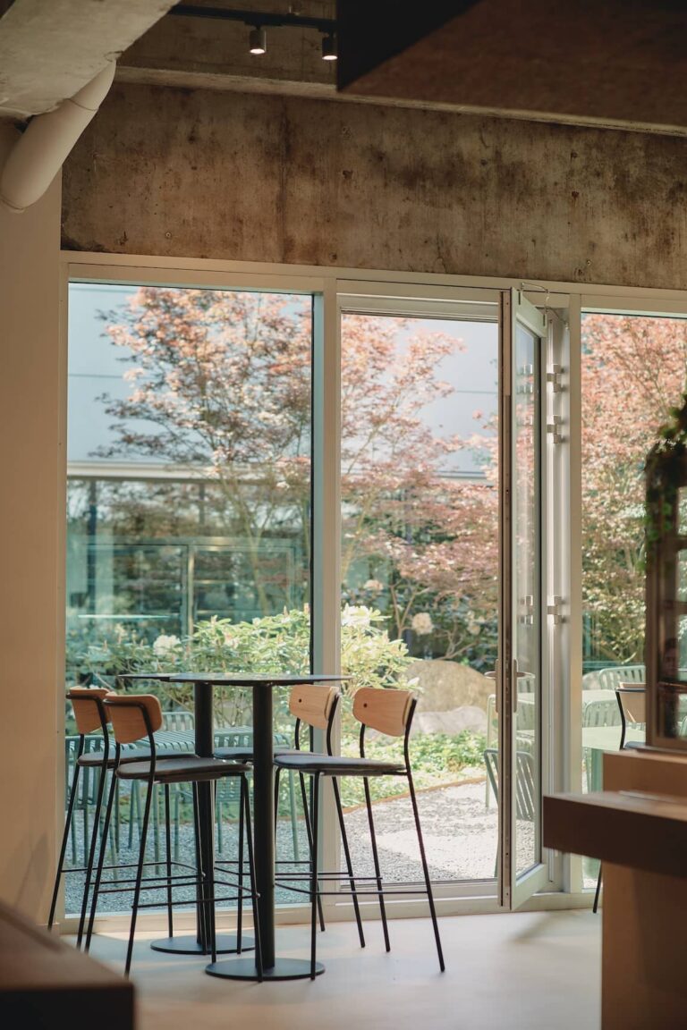High tables and stools near an open glass door, connecting the indoor kitchen area with a garden outside, blending natural light and soft greenery.