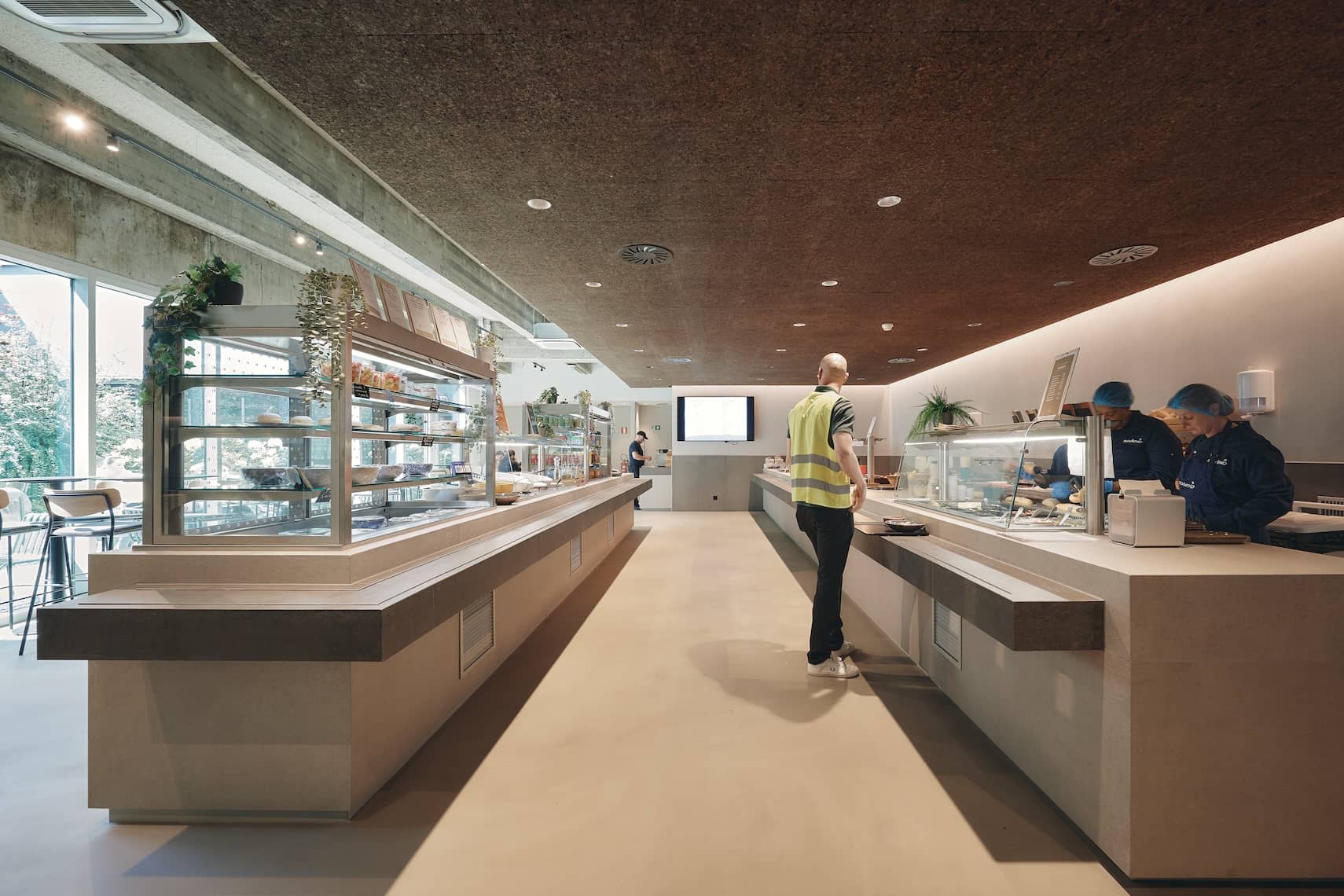 Staff members serve lunch behind a long modern kitchen counter with clean lines, natural materials, and cork ceiling panels.