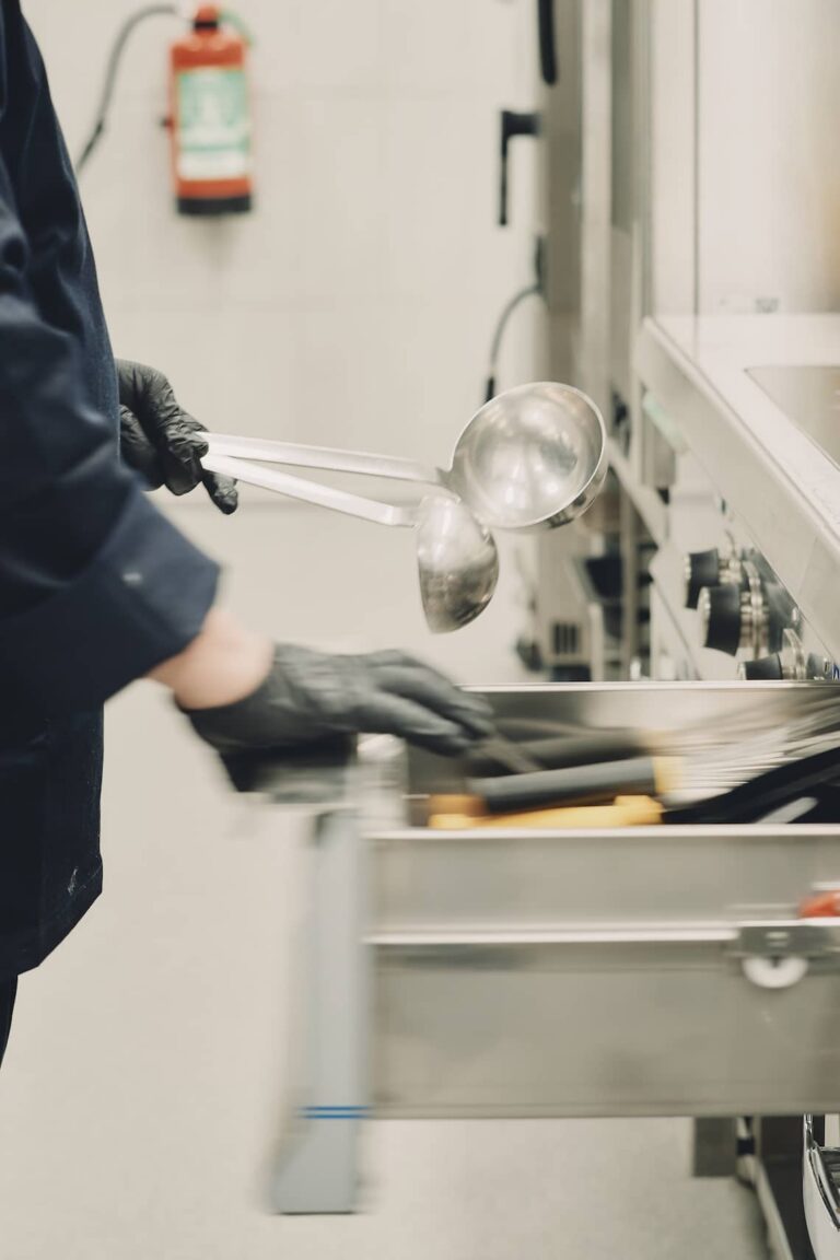 Chef with gloves handling stainless steel ladles in an industrial kitchen drawer