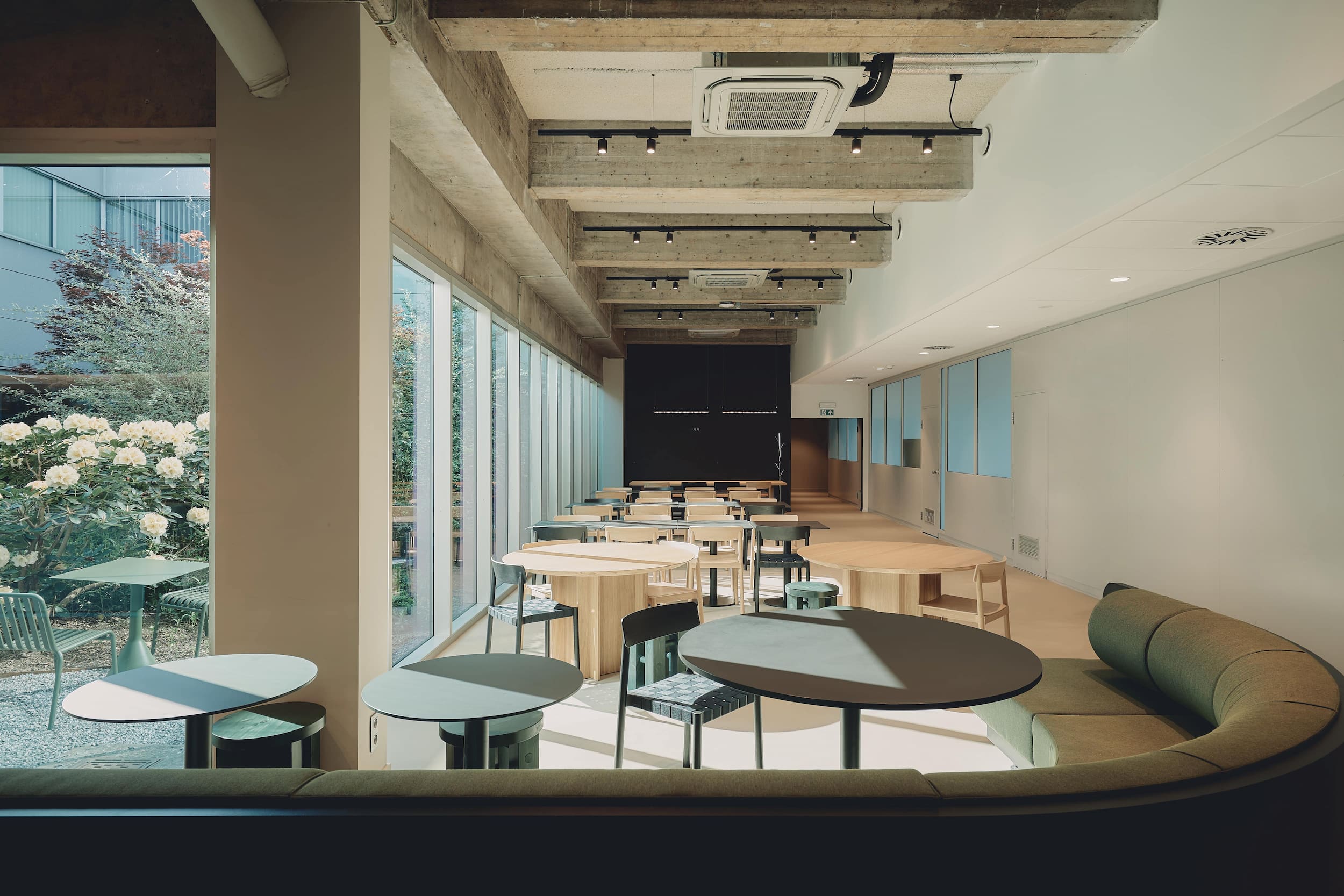 Contemporary dining area with custom circular and oval tables, green upholstered bench seating, and floor-to-ceiling windows overlooking a lush outdoor garden