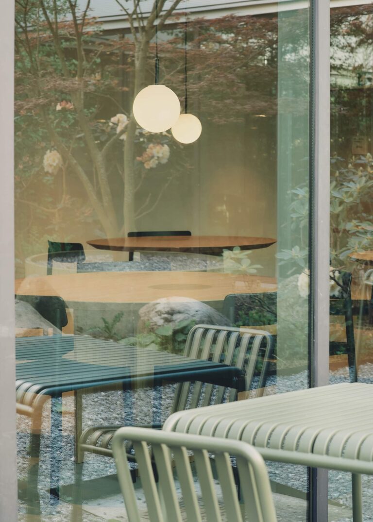 Close-up view through glass showing overlapping reflections of outdoor garden furniture, indoor round tables, pendant lights, and flowering trees
