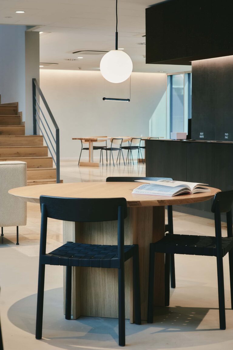 Detail of a round wooden table with black woven chairs, an open book, and a globe pendant light in a modern, minimalist interior with warm lighting and natural materials