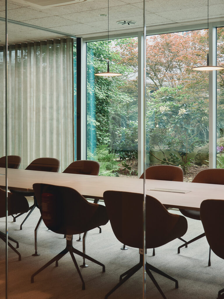 Modern meeting room with a long wooden table, swivel chairs, pendant lights, and floor-to-ceiling windows offering a view of a lush garden with trees and plants